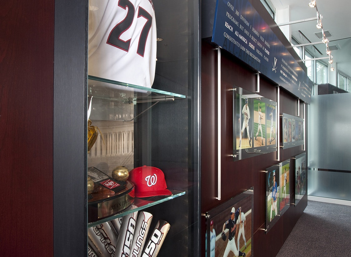 The opposite wall holds a mixed media exhibit of display cases. Glass cases hold the program’s most prestigious championship trophies and player artifacts along with recognition of those players who have made it to the MLB. This room physically distills the success of the program over time and inspires incoming players to consider what their future might hold at UVA as well. 